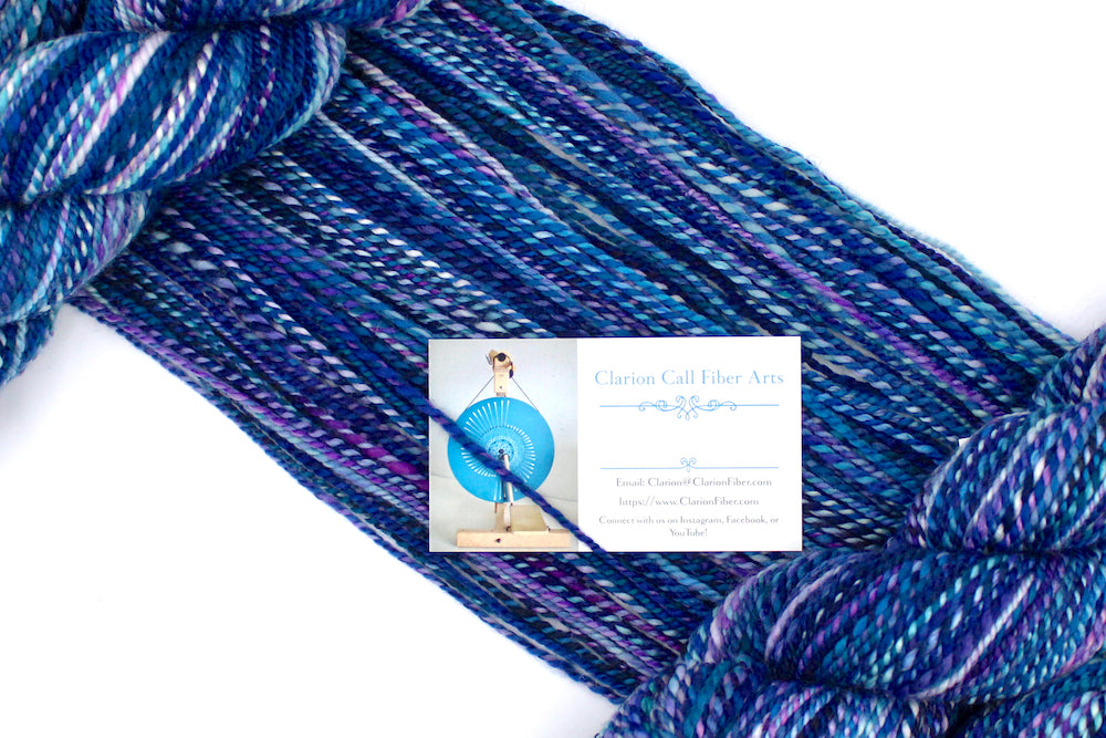 A one of a kind, Dk Weight, hand dyed variegated skein of multicolored Blue,and Purple self-striping Hand spun Yarn, made of 100% Hand Dyed Merino Wool, draped diagonally across the frame, so you can really see the color play. 