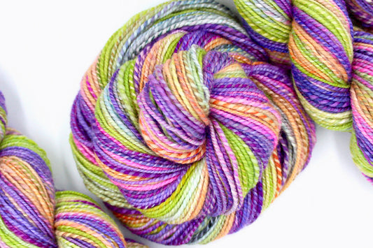 A one of a kind, hand dyed gradient skein of multicolored Purple, Neon Pink, Orange, Yellow Neon Green, and Light Blue self-striping wool Yarn coiled attractively in the center of the frame. 