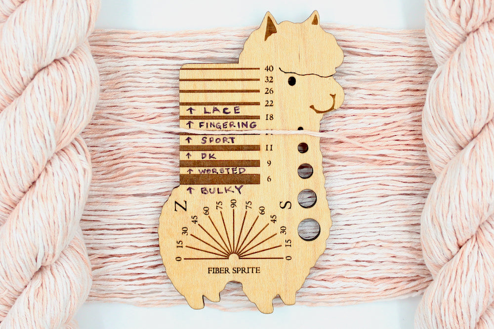 A skein of Vegan, Variegated Pink and Cream, 100% Cotton, Sport Weight Yarn recycled by hand from unwanted sweaters draped over the center of the frame with one strand on top of a Llama shaped Yarn weight tool, to better illustrate the thickness of the yarn. 