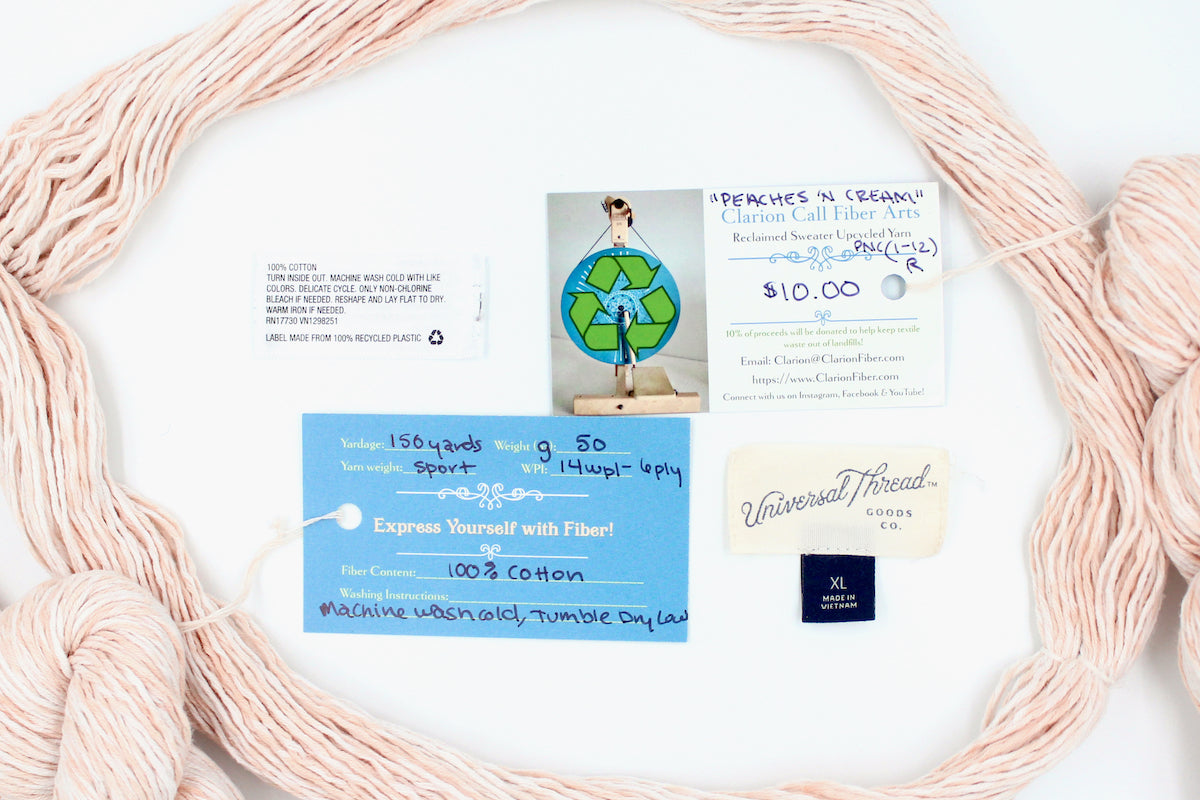 A skein of Vegan, Variegated Pink and Cream, 100% Cotton, Sport Weight yarn encircling the sweater labels it was recycled from, as well as the hand written yarn tags detailing the specs of this recycled yarn to help you make your next project! 