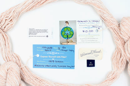 A skein of Vegan, Variegated Pink and Cream, 100% Cotton, Sport Weight yarn encircling the sweater labels it was recycled from, as well as the hand written yarn tags detailing the specs of this recycled yarn to help you make your next project! 
