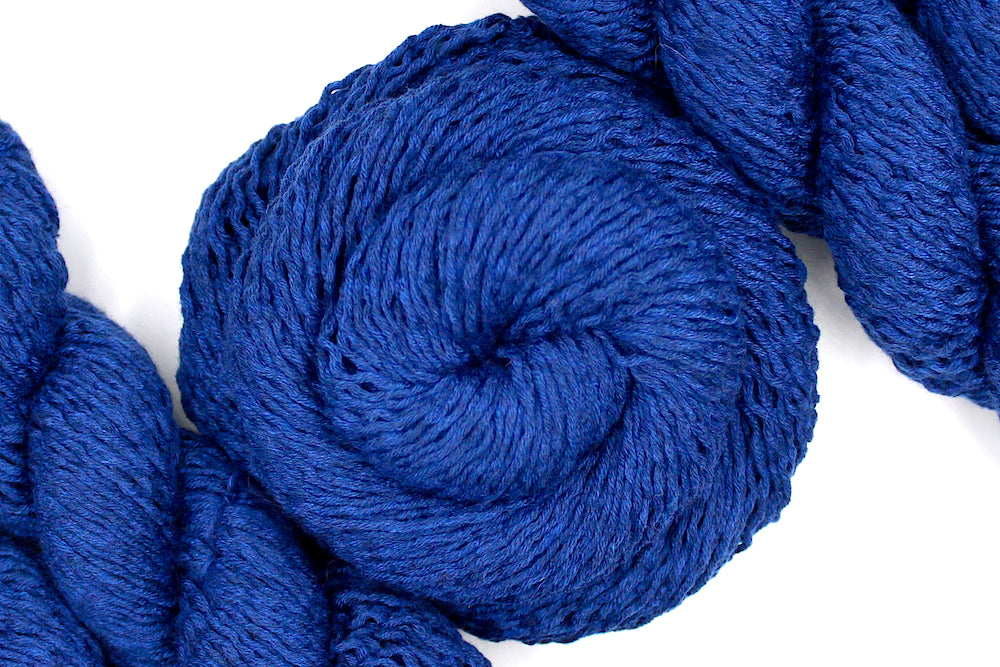 A skein of Vegan, Purplish Blue, 85% Polyester, 15% Nylon, Sport Weight Yarn recycled by hand from unwanted sweaters swirled attractively in the center of the frame. 