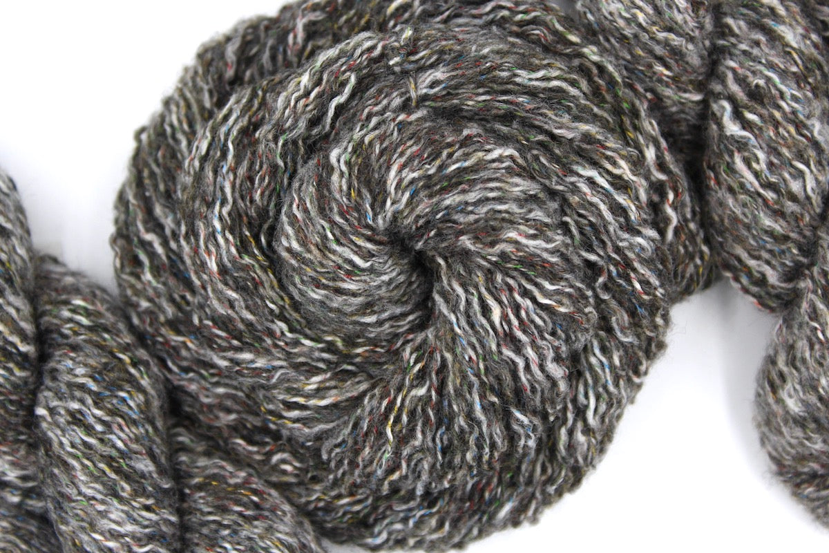 A skein of Vegan, Greyish Brown Variegated, Cotton/ Acrylic/ Polyester, DK weight Yarn recycled by hand from unwanted sweaters swirled attractively in the center of the frame. 