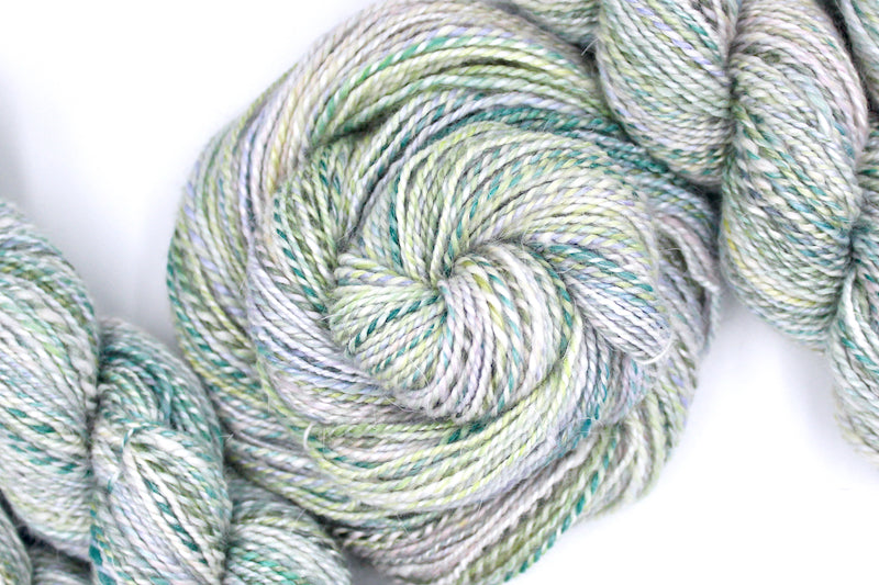 A one of a kind, hand dyed variegated skein of multicolored pastel Green, Lavender, and Pink self-striping wool Yarn coiled attractively in the center of the frame. 