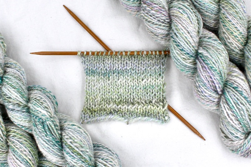 A sample swatch knitted from a one of a kind, hand dyed variegated skein of multicolored pastel Green, Lavender, and Pink self-striping wool Yarn. 