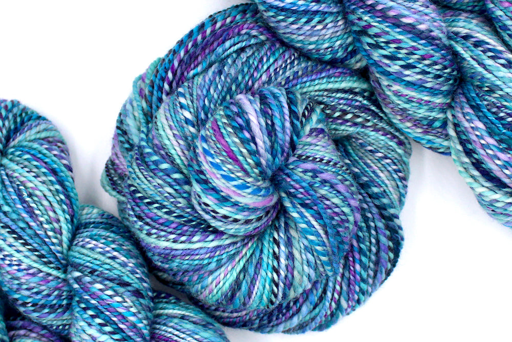 A one of a kind, Dk Weight, hand dyed variegated skein of multicolored Blue, Pink, and Purple self-striping Hand spun Yarn, made of 100% Hand Dyed Superwash Merino Wool and coiled attractively in the center of the frame. 