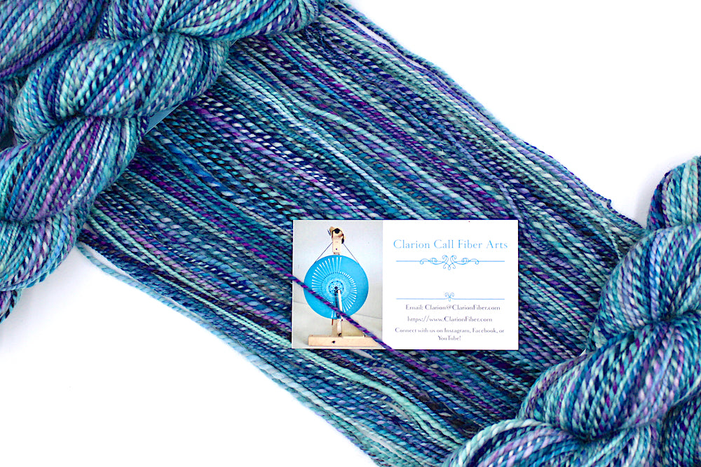 A one of a kind, Dk Weight, hand dyed variegated skein of multicolored Blue, Pink, and Purple self-striping Hand spun Yarn, made of 100% Hand Dyed Superwash Merino Wool, draped diagonally across the frame, so you can really see the color play. 