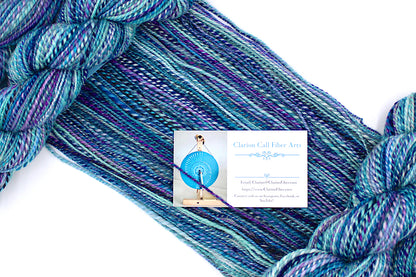 A one of a kind, Dk Weight, hand dyed variegated skein of multicolored Blue, Pink, and Purple self-striping Hand spun Yarn, made of 100% Hand Dyed Superwash Merino Wool, draped diagonally across the frame, so you can really see the color play. 