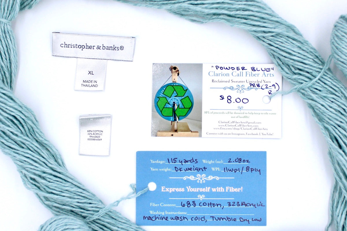 A skein of Vegan, Pastel Baby Blue, Cotton/ Acrylic, Dk Weight yarn encircling the sweater labels it was recycled from, as well as the hand written yarn tags detailing the specs of this upcycled yarn to help you make your next project! 