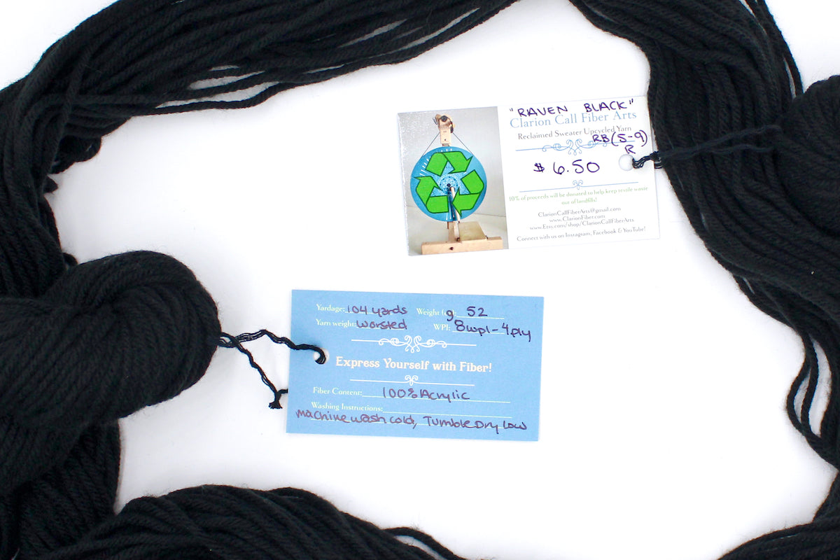 A skein of Vegan, Black, 100% Acrylic, Worsted Weight yarn encircling the sweater labels it was recycled from, as well as the hand written yarn tags detailing the specs of this recycled yarn to help you make your next project! 