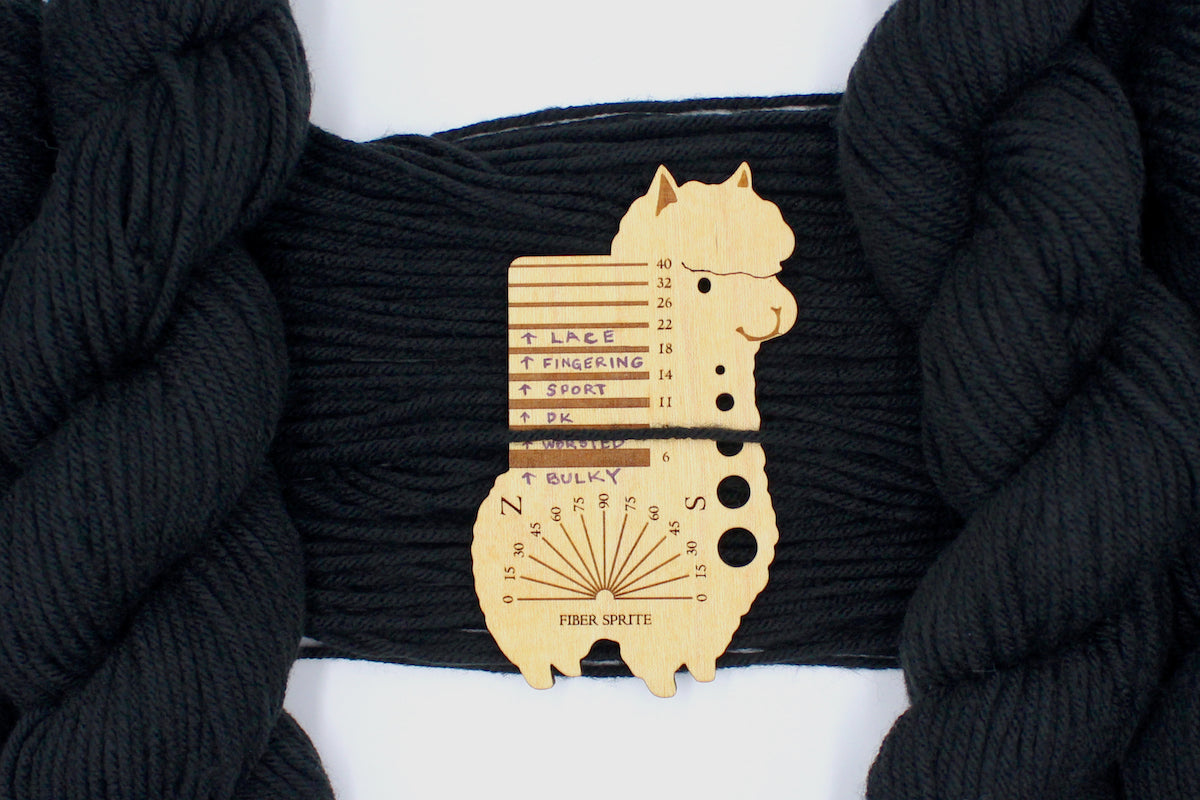 A skein of Vegan, Black, 100% Acrylic, Worsted Weight Yarn recycled by hand from unwanted sweaters draped over the center of the frame with one strand on top of a Llama shaped Yarn weight tool, to better illustrate the thickness of the yarn. 