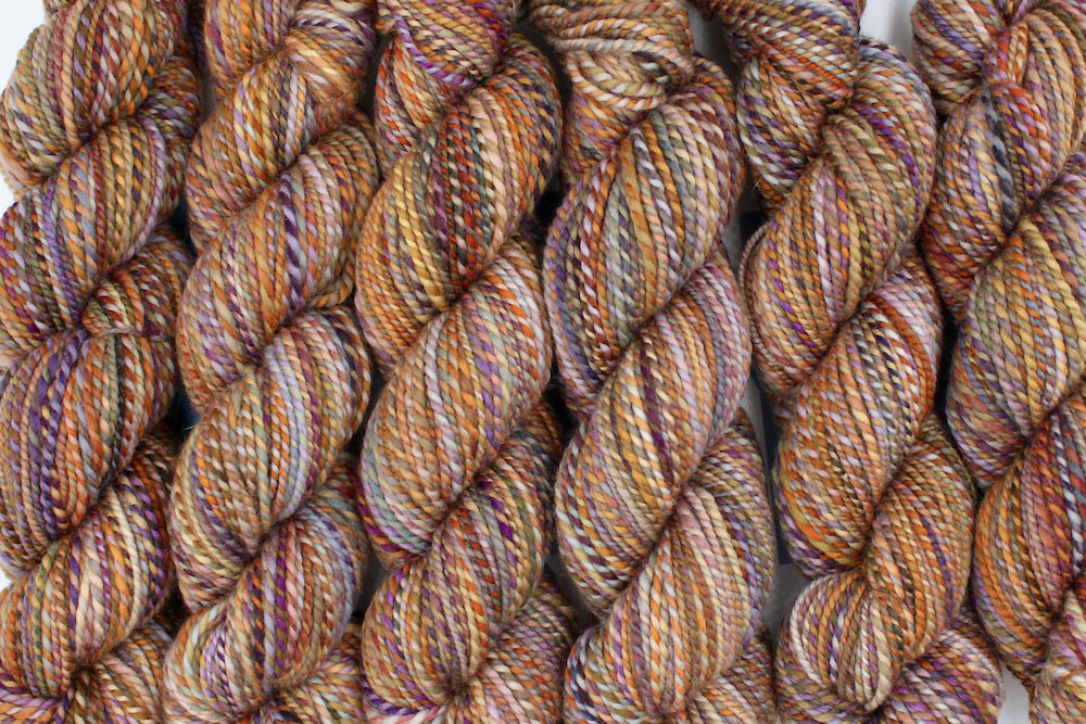 Six one of a kind, Worsted weight, hand dyed variegated skein of multicolored Orange, Yellow, Pink, Purple, Olive Green, and Blue self-striping wool Yarn lined up side by side. 