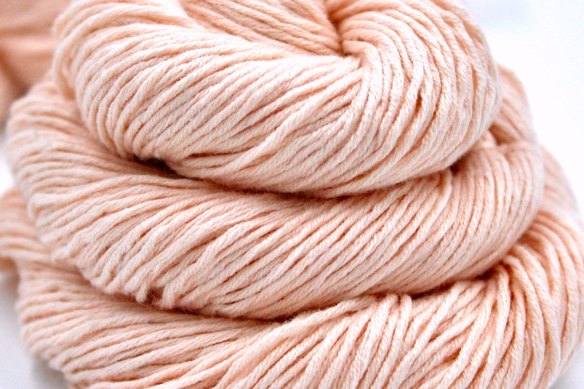 A close up shot of a skein of Vegan, Light Pastel Peach/ Pink, Cotton/ Rayon/ Nylon, Sport weight Yarn recycled by hand from unwanted sweaters beautifully coiled in the center of the frame. 