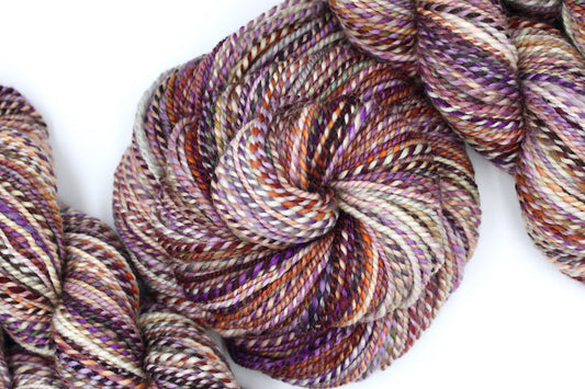 A one of a kind, hand dyed Variegated skein of multicolored Maroon, Orange, Gold, Purple, and Green self-striping wool Yarn coiled attractively in the center of the frame. 