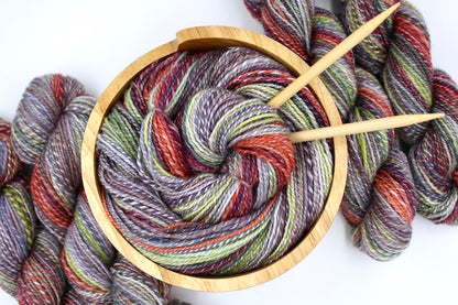 A one of a kind, hand dyed gradient skein of multicolored Grey, Lime Green, Olive Green, Maroon, and Bright Peach self-striping wool Yarn, in a yarn bowl with knitting needles, ready to be made into something beautiful! 