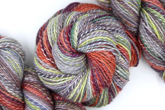 A one of a kind, hand dyed gradient skein of multicolored Grey, Lime Green, Olive Green, Maroon, and Bright Peach self-striping wool Yarn coiled attractively in the center of the frame. 