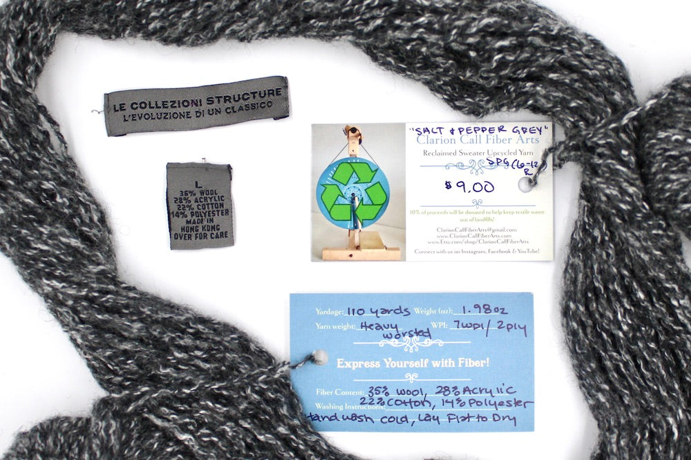 A skein of Heathered Charcoal Grey and White, Wool/ Acrylic/ Cotton/ Polyester, Heavy Worsted Weight yarn encircling the sweater labels it was recycled from, as well as the hand written yarn tags detailing the specs of this upcycled yarn to help you make your next project! 
