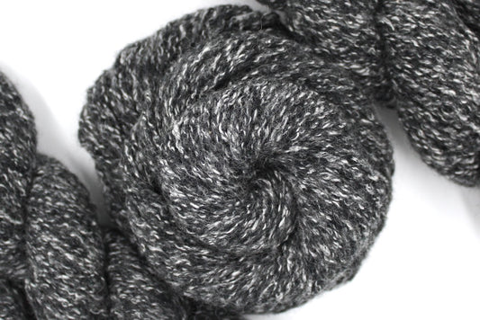 A skein of Heathered Charcoal Grey and White, Wool/ Acrylic/ Cotton/ Polyester, Heavy Worsted weight Yarn recycled by hand from unwanted sweaters swirled attractively in the center of the frame. 