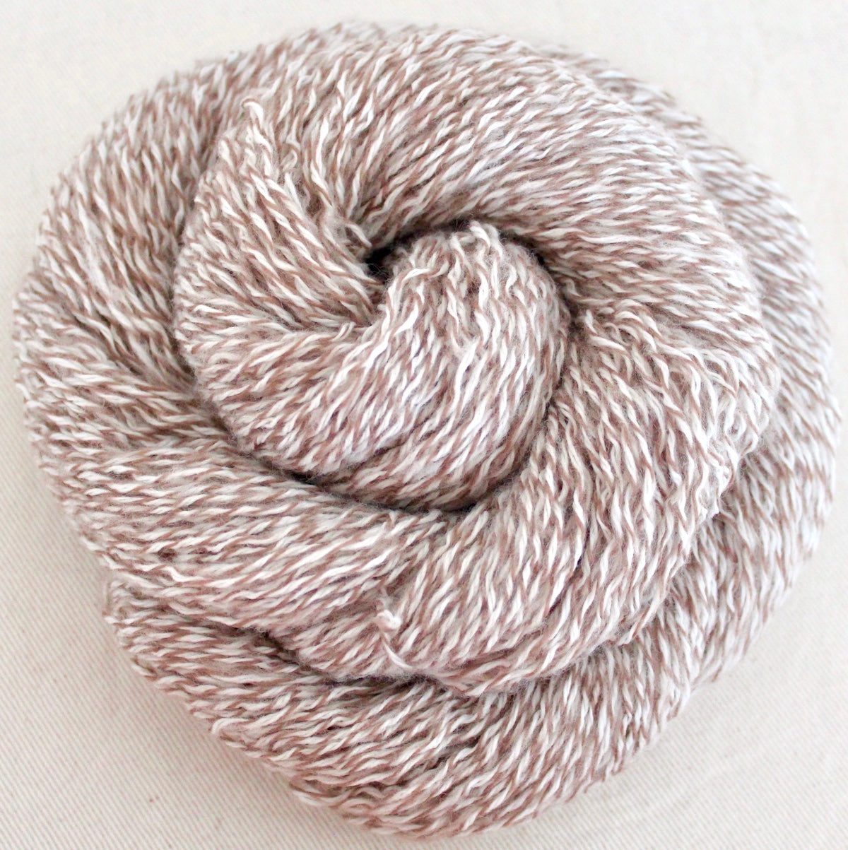 A skein of Vegan, Variegated Tan and White, 100% Acrylic, Sport weight Yarn recycled by hand from unwanted sweaters swirled attractively in the center of the frame. 