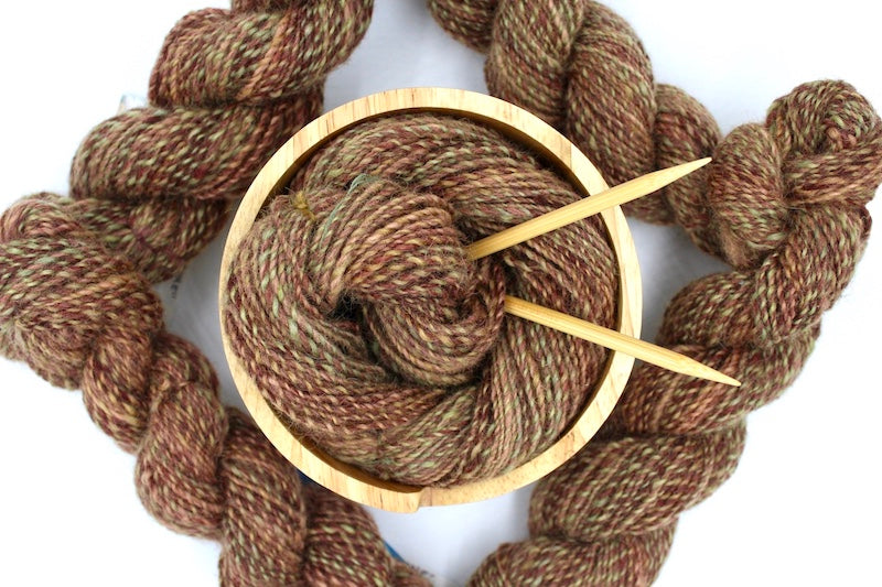 A one of a kind, hand dyed variegated skein of multicolored Reddish Brown, Green, Taupe, and Gold self-striping wool Yarn, in a yarn bowl with knitting needles, ready to be made into something beautiful! 