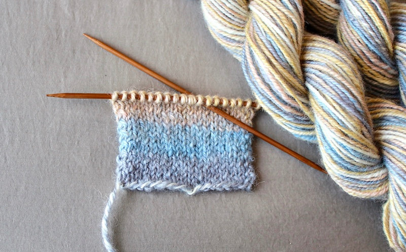 A sample swatch knitted from a one of a kind, hand dyed multicolored Grey, Baby Blue, Pastel Pink, Yellow, and White self-striping wool Yarn. 