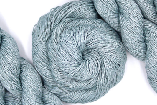 A skein of Vegan, Light Grayish Blue, 100% Linen, Fingering weight Yarn recycled by hand from unwanted sweaters swirled attractively in the center of the frame. 