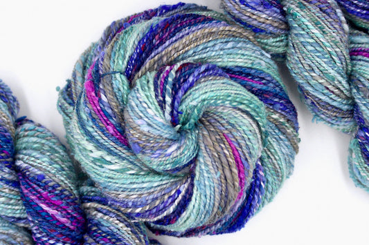 A one of a kind, hand dyed gradient skein of multicolored Seafoam Green, Steel Blue, Grey, Lavender, Deep Violet, and Fuchsia self-striping wool Yarn coiled attractively in the center of the frame. 
