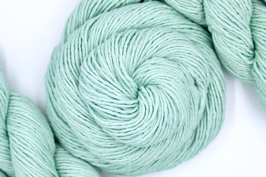 A skein of Vegan, Seafoam/ Aquamarine Blueish Green, 100% Cotton, Dk weight Yarn recycled by hand from unwanted sweaters swirled attractively in the center of the frame. 