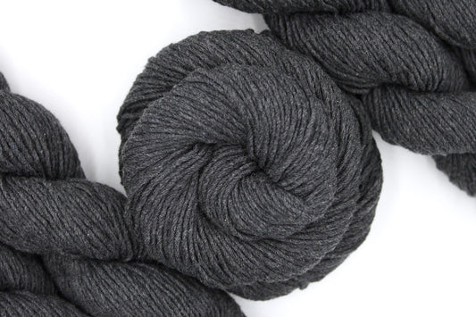 A skein of Vegan, Dark Heathered Charcoal Grey, 100% Cotton, Worsted weight Yarn recycled by hand from unwanted sweaters swirled attractively in the center of the frame. 