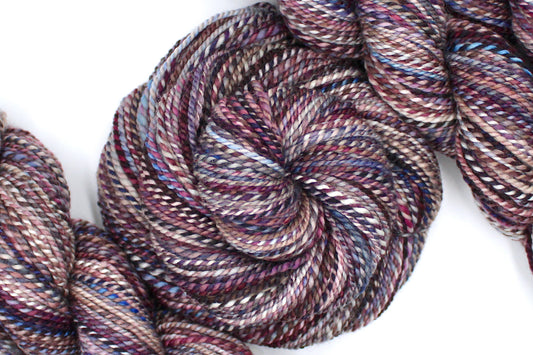 A one of a kind, Dk Weight, hand dyed variegated skein of multicolored Maroon, Burgandy, Purple, Grey, and Blue self-striping 100% Superwash Merino wool Yarn coiled attractively in the center of the frame. 