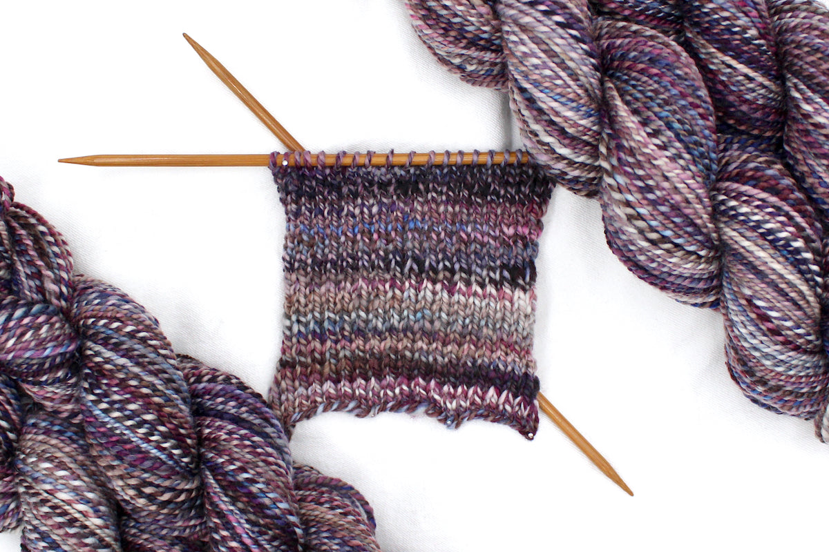 A sample swatch knitted on size 5 knitting needles from a one of a kind, Dk Weight, hand dyed variegated skein of multicolored Maroon, Burgandy, Purple, Grey, and Blue self-striping 100% Superwash Merino wool Yarn. 