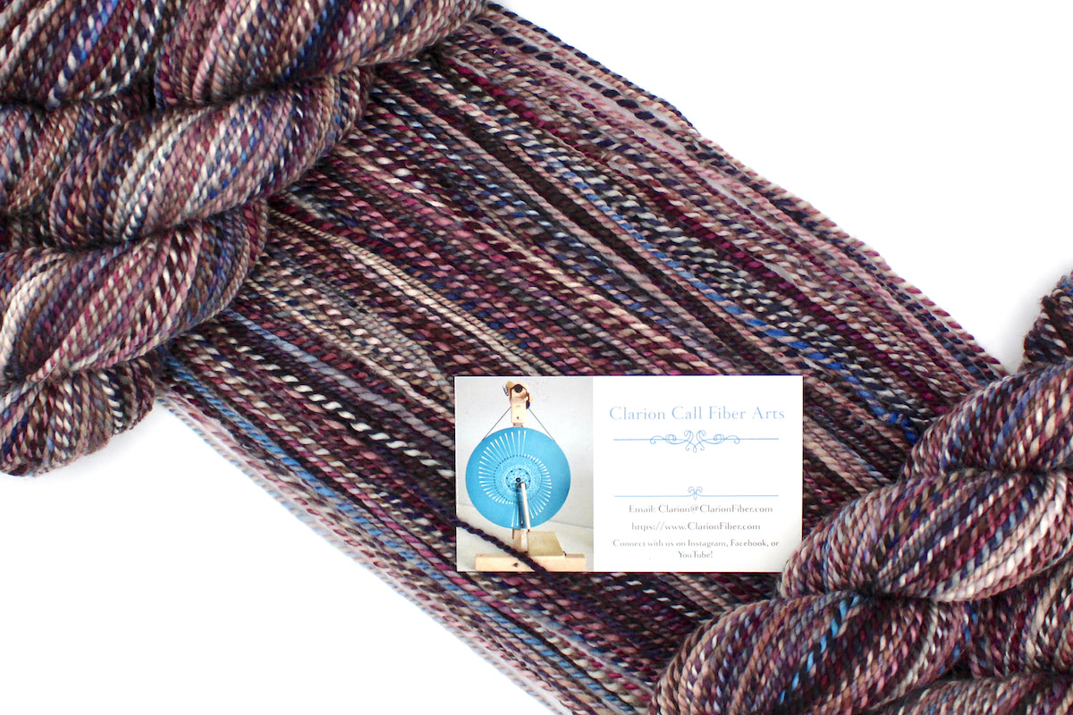 A one of a kind, Dk Weight, hand dyed variegated skein of multicolored Maroon, Burgandy, Purple, Grey, and Blue self-striping 100% Superwash Merino wool Yarn draped diagonally across the frame, so you can really see the color play. 
