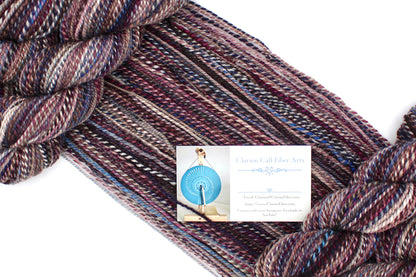 A one of a kind, Dk Weight, hand dyed variegated skein of multicolored Maroon, Burgandy, Purple, Grey, and Blue self-striping 100% Superwash Merino wool Yarn draped diagonally across the frame, so you can really see the color play. 