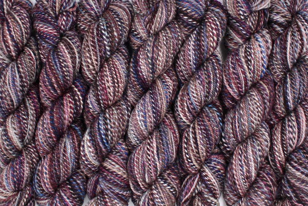Six one of a kind, Dk Weight, hand dyed variegated skein of multicolored Maroon, Burgandy, Purple, Grey, and Blue self-striping 100% Superwash Merino wool Yarn lined up side by side. 