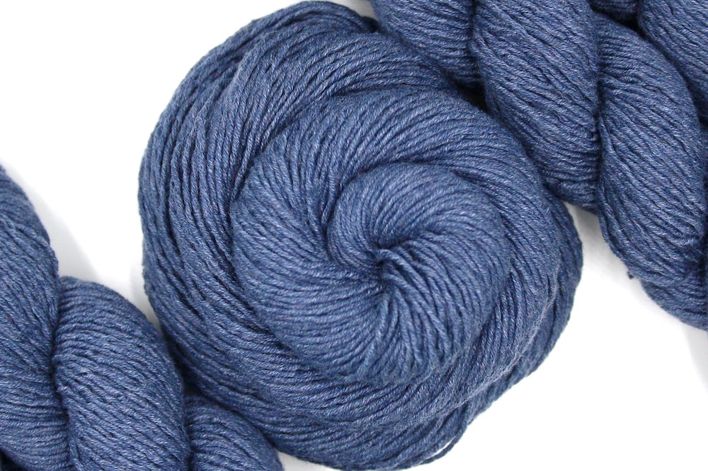 A skein of Vegan, Heathered Slate Grayish Blue, Cotton/ Acrylic, Sport weight Yarn recycled by hand from unwanted sweaters swirled attractively in the center of the frame. 