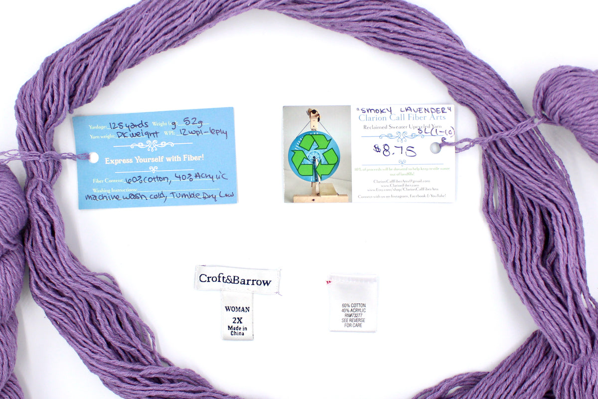 A skein of Vegan, Dusky Lavender, 60% Cotton, 40% Acrylic, DK Weight yarn encircling the sweater labels it was recycled from, as well as the hand written yarn tags detailing the specs of this recycled yarn to help you make your next project! 