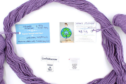 A skein of Vegan, Dusky Lavender, 60% Cotton, 40% Acrylic, DK Weight yarn encircling the sweater labels it was recycled from, as well as the hand written yarn tags detailing the specs of this recycled yarn to help you make your next project! 