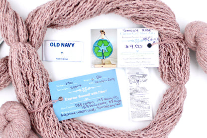 A skein of Vegan, Dusky Pink, Cotton, Polyester, Acrylic, Nylon, Sport Weight yarn encircling the sweater labels it was recycled from, as well as the hand written yarn tags detailing the specs of this recycled yarn to help you make your next project! 