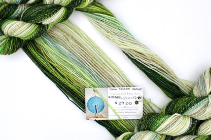 A one of a kind, hand dyed gradient skein of multicolored Evergreen, Olive, Lime Green, Seafoam Green, Light Grayish Brown, and Cream self-striping wool Yarn draped diagonally across the frame, so you can really see the color play. 