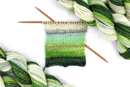 A sample swatch knitted from a one of a kind, hand dyed gradient skein of multicolored Evergreen, Olive, Lime Green, Seafoam Green, Light Grayish Brown, and Cream self-striping wool Yarn. 