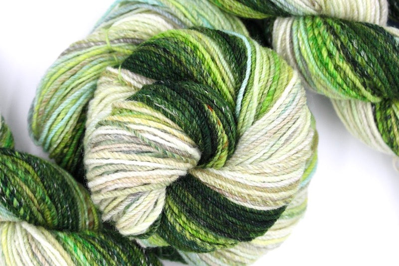 A one of a kind, hand dyed gradient skein of multicolored Evergreen, Olive, Lime Green, Seafoam Green, Light Grayish Brown, and Cream self-striping wool Yarn coiled attractively in the center of the frame. 
