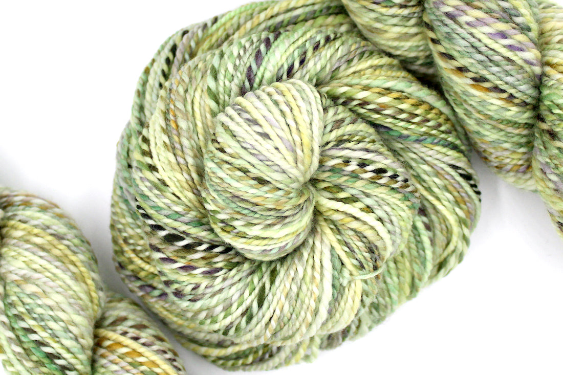 A one of a kind Dk Weight, hand dyed variegated skein of multicolored Green, Yellow, Lavender, and Tan self-striping Hand spun Yarn, made of 100% Hand Dyed Superwash Merino Wool; coiled attractively in the center of the frame. 