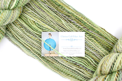 A one of a kind, Dk Weight, hand dyed variegated skein of multicolored Green, Yellow, Lavender, and Tan self-striping Hand spun Yarn, made of 100% Hand Dyed Superwash Merino Wool, draped diagonally across the frame, so you can really see the color play. 