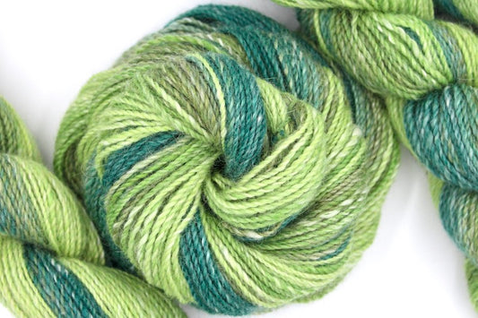 A one of a kind, hand dyed gradient skein of multicolored Dark Jungle Green, Olive, Lime Green, and Light Spring Green self-striping wool Yarn coiled attractively in the center of the frame. 