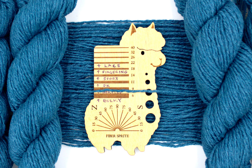 A skein of Vegan, Dark Greenish Teal, Cotten blend, Worsted Weight Yarn recycled by hand from unwanted sweaters draped over the center of the frame with one strand on top of a Llama shaped Yarn weight tool, to better illustrate the thickness of the yarn. 