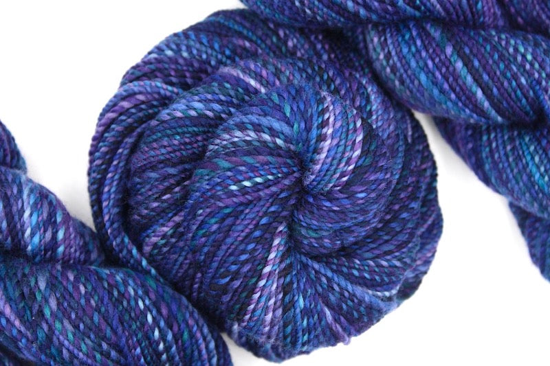 A one of a kind, hand dyed variegated skein of multicolored Navy Blue, Plum, Sapphire Blue, Teal and Lavender self-striping wool Yarn coiled attractively in the center of the frame. 