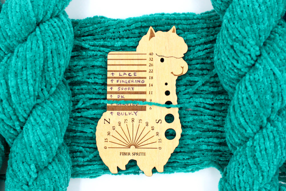 A skein of Vegan, Bright Teal, 100% Acrylic, Worsted Weight Chenille Yarn recycled by hand from unwanted sweaters draped over the center of the frame with one strand on top of a Llama shaped Yarn weight tool, to better illustrate the thickness of the yarn. 