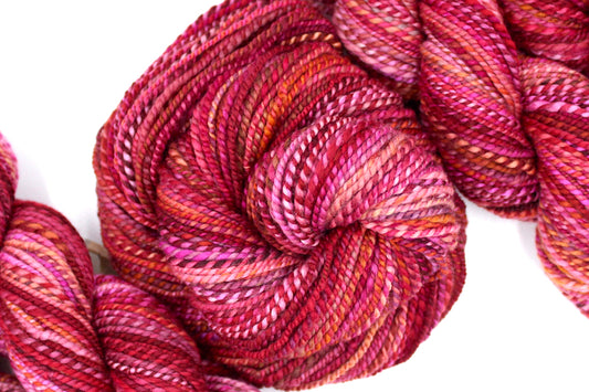 A one of a kind, hand dyed variegated skein of multicolored Red, Pink, and Orange self-striping wool Yarn coiled attractively in the center of the frame.