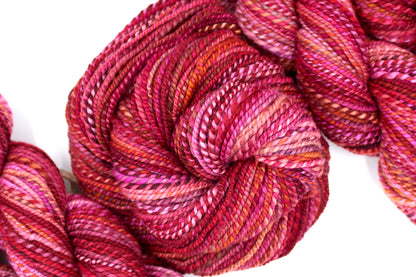 A one of a kind, hand dyed variegated skein of multicolored Red, Pink, and Orange self-striping wool Yarn coiled attractively in the center of the frame. 