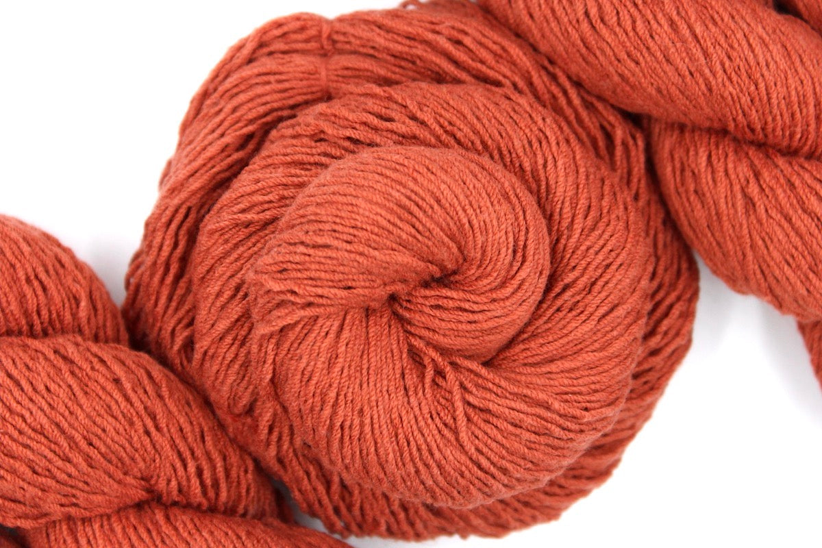 A skein of Vegan, Rusty Red/ Terracotta Orange, Cotton/ Acrylic, Fingering weight Yarn recycled by hand from unwanted sweaters swirled attractively in the center of the frame. 